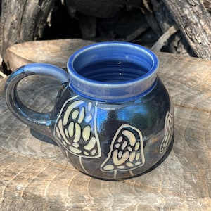 May include: A handcrafted ceramic mug with a dark blue exterior and a lighter blue interior. The mug showcases a whimsical design of white and cream-coloured mushrooms. The handle and rim are also blue, creating a cohesive and artistic look.
