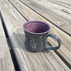 May include: A white ceramic mug with a black handle and the text "But First, Coffee" in a bold, black font. The mug is filled with a dark liquid, likely coffee, and sits on a light-colored surface.