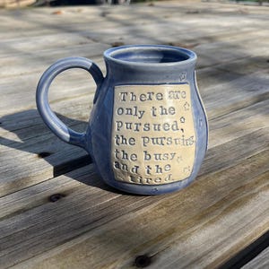 May include: A blue ceramic mug with a curved handle. The mug features a rectangular cream-colored panel with the quote "There are only the pursued, the pursuing, the busy and the tired." Small star designs are scattered around the panel.