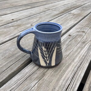 May include: A handmade ceramic mug with a blue handle and rim. The mug's body is dark blue with a wheat stalk design in white. The mug is sitting on a weathered wooden surface.