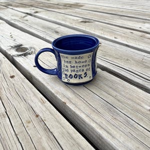 May include: A blue ceramic mug with a handle, featuring the text "She made her home in between the pages of BOOKS." The mug has a white panel with the text and star designs. The mug is sitting on a wooden surface.