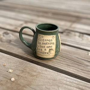 May include: A handcrafted, dark green ceramic mug with a curved handle. The mug features a cream-colored rectangular label with the quote, "The distance is nothing when one has a motive." Decorative leaf patterns are etched on the mug's surface.