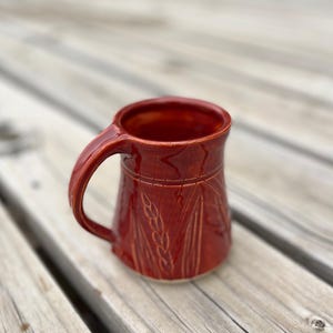 Peut inclure: Une tasse en céramique rouge faite à la main avec une grande anse. La tasse présente des motifs de tiges de blé gravés et des lignes horizontales sur le dessus. La tasse est posée sur une surface en bois.