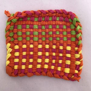 May include: A square, woven hot pad in a vibrant mix of colors. The pad features a repeating pattern of orange, pink, green, and yellow fabric strips. The edges are braided with orange, pink, and green fabric, and a loop is attached for hanging.