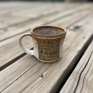 May include: A white ceramic mug with a brown glaze and a gold rectangle on the side. The rectangle has the text "BOOKS may well be the only true MAGIC Alice Hoffman" in black lettering.