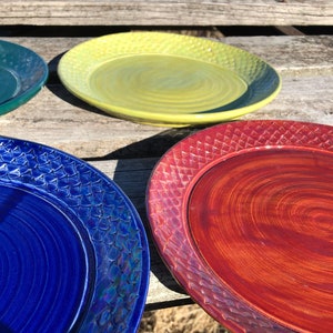 May include: Four ceramic plates in different colors: green, yellow, blue, and red. The plates have a textured rim and a smooth center.