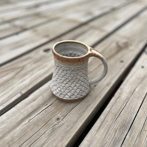 May include: A white ceramic mug with a textured, geometric pattern and a brown rim. The mug has a handle and is sitting on a wooden surface.