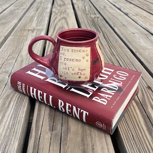 May include: A burgundy ceramic mug with a curved handle, featuring the text "You rescue me, I rescue you. That's how this works." resting on a hardcover book titled "Hell Bent" by Leigh Bardugo.