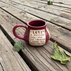 May include: A burgundy ceramic mug with a white handle and a white inscription that reads "LOVE is peculiar magic, she said. Because it makes you strong but also delicate."