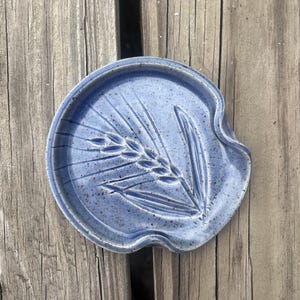 May include: A round, blue ceramic spoon rest with a speckled finish and a debossed wheat stalk design. The piece features a curved edge indentation on the right side, designed to hold the handle of a kitchen utensil securely in place.