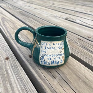 May include: A dark green ceramic mug with a curved handle. The mug features the text "Let's have a toast. To the incompetence of our ENEMIES" in a light blue font. Decorative elements include stars and a mushroom.