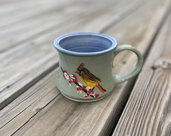 May include: A handmade ceramic mug with a painted cardinal bird design. The mug features a light green exterior, a blue interior, and a curved handle. The bird is perched on a branch with red berries. The mug is sitting on a wooden surface.