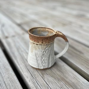 May include: A handmade ceramic mug with a wheat stalk design. The mug features a light beige body with a brown rim and handle. The handle is curved and the mug is approximately 4 inches tall.