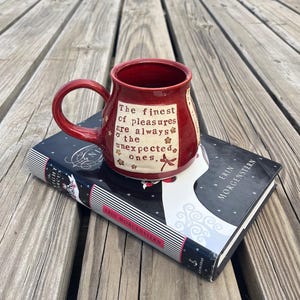 May include: A red ceramic mug with a curved handle, resting on a book. The mug features the text "The finest of pleasures are always the unexpected ones." The book has a black cover with white and red accents.