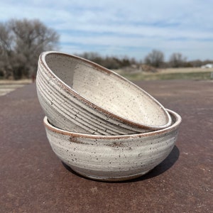 May include: Two white ceramic bowls with brown rims are stacked on top of each other. The bowls have a speckled glaze and a slightly textured surface.