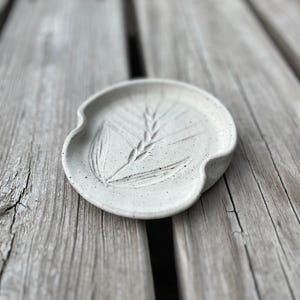 May include: A speckled, off-white ceramic spoon rest with a wheat stalk design. The spoon rest has a curved shape with a notch on one side. The wheat stalk is etched into the surface.