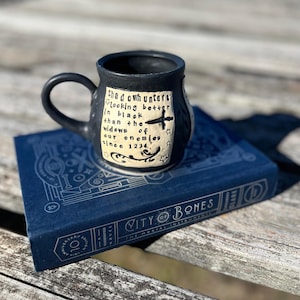 May include: Black ceramic mug with a white design featuring a quote from the book series "Shadowhunters" by Cassandra Clare. The quote reads "Shadowhunters looking better in black than the widows of our enemies since 1234". The mug is sitting on a blue book with the title "City of Bones" and the subtitle "The Mortal Instruments".