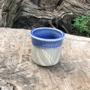 May include: A small, white ceramic mug with a blue rim and a design of wheat stalks. The mug is sitting on a wooden surface.