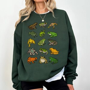 May include: Dark green sweatshirt featuring a collection of illustrated frogs in various colors and poses. The frogs are arranged in a grid pattern across the front of the sweatshirt. The sweatshirt has a crew neck and long sleeves.