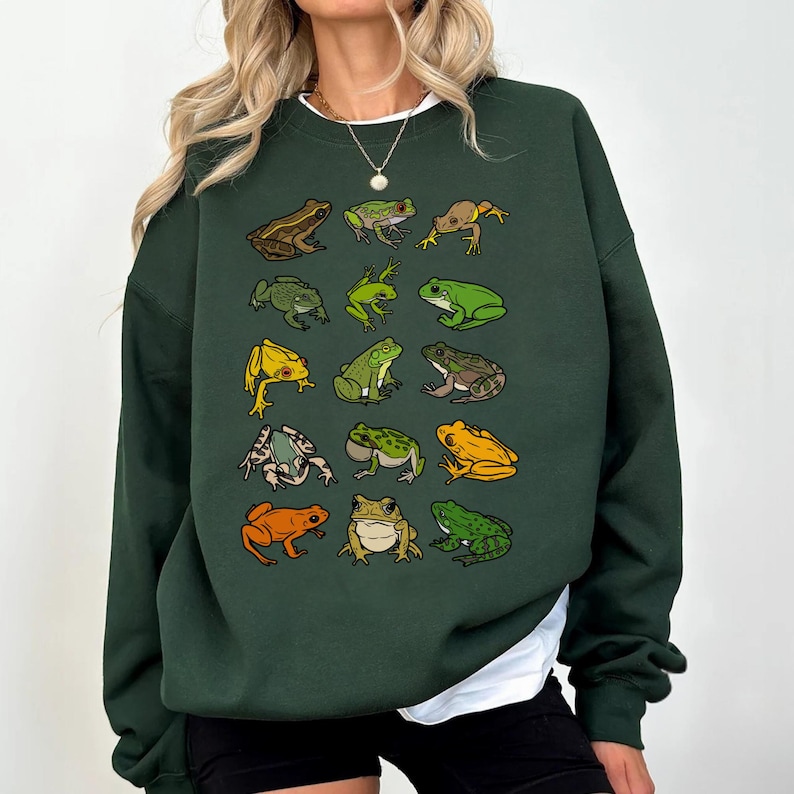 May include: Dark green sweatshirt featuring a collection of illustrated frogs in various colors and poses. The frogs are arranged in a grid pattern across the front of the sweatshirt. The sweatshirt has a crew neck and long sleeves.