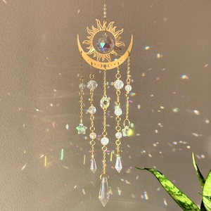 Puede incluir: Un atrapasoles de tem&aacute;tica celestial con un dise&ntilde;o de sol y luna creciente de color dorado. Cuentas y colgantes de cristal transparente cuelgan de la luna, con una gran bola de cristal en el centro. El atrapasoles est&aacute; suspendido por una delicada cadena.