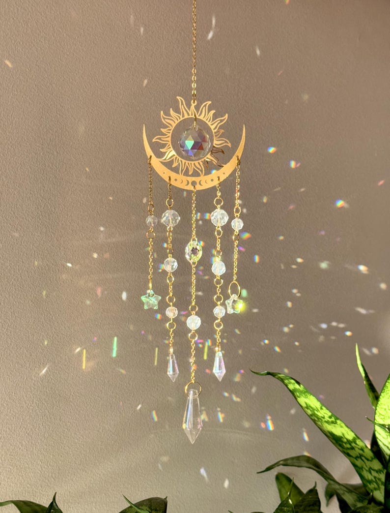 Puede incluir: Un atrapasoles de tem&aacute;tica celestial con un dise&ntilde;o de sol y luna creciente de color dorado. Cuentas y colgantes de cristal transparente cuelgan de la luna, con una gran bola de cristal en el centro. El atrapasoles est&aacute; suspendido por una delicada cadena.