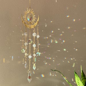 Puede incluir: Un atrapasoles de tem&aacute;tica celestial con un dise&ntilde;o de sol y luna creciente en color dorado. Cuentas de cristal transparente y adornos en forma de estrella cuelgan del sol y la luna. El atrapasoles est&aacute; suspendido por una delicada cadena, creando un efecto decorativo y et&eacute;reo.