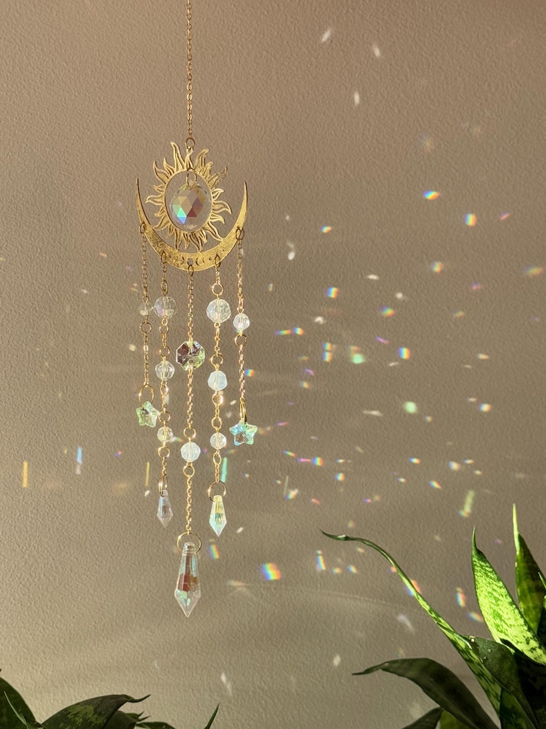 Puede incluir: Un atrapasoles de tem&aacute;tica celestial con un dise&ntilde;o de sol y luna creciente en color dorado. Cuentas de cristal transparente y adornos en forma de estrella cuelgan del sol y la luna. El atrapasoles est&aacute; suspendido por una delicada cadena, creando un efecto decorativo y et&eacute;reo.