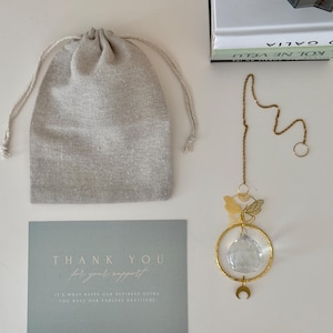 Puede incluir: Un atrapasoles de cristal dorado con un dise&ntilde;o de mariposa y luna, colgando de una cadena. Incluye una bolsa con cord&oacute;n y una tarjeta con el texto "THANK YOU for your support".