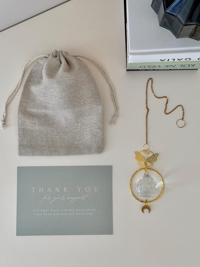 Puede incluir: Un atrapasoles de cristal dorado con un dise&ntilde;o de mariposa y luna, colgando de una cadena. Incluye una bolsa con cord&oacute;n y una tarjeta con el texto "THANK YOU for your support".