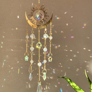 Puede incluir: Adorno colgante dorado con forma de sol y luna creciente, con cuentas y prismas de cristal transparente. El sol tiene rayos detallados, y la luna acuna un gran cristal. El adorno cuelga de una cadena dorada.