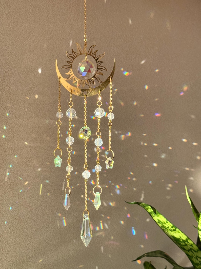 Puede incluir: Adorno colgante dorado con forma de sol y luna creciente, con cuentas y prismas de cristal transparente. El sol tiene rayos detallados, y la luna acuna un gran cristal. El adorno cuelga de una cadena dorada.