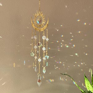 Puede incluir: Un atrapasoles dorado con un dise&ntilde;o de sol y media luna. El atrapasoles presenta un dise&ntilde;o de sol con un cristal en el centro y cuentas de cristal colgantes. El atrapasoles est&aacute; suspendido por una cadena.