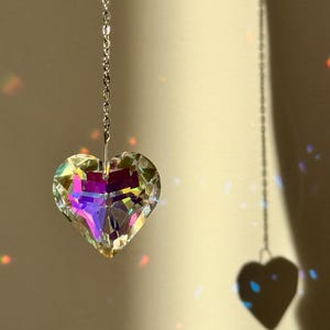 Op de afbeelding: Een kristallen hartvormig prisma dat aan een zilveren ketting hangt. Het prisma werpt regenbogen op de muur erachter.