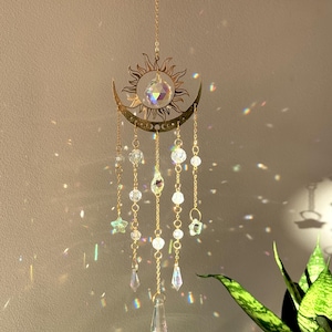 Puede incluir: Un atrapasoles de tem&aacute;tica celestial con un dise&ntilde;o de sol y luna creciente dorados. Cuentas de cristal transparente y adornos en forma de estrella cuelgan de la luna. El atrapasoles est&aacute; suspendido por una delicada cadena, creando un efecto decorativo y et&eacute;reo.
