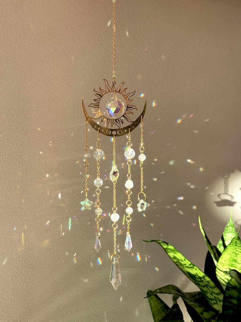 Puede incluir: Un atrapasoles de tem&aacute;tica celestial con un dise&ntilde;o de sol y luna creciente dorados. Cuentas de cristal transparente y adornos en forma de estrella cuelgan de la luna. El atrapasoles est&aacute; suspendido por una delicada cadena, creando un efecto decorativo y et&eacute;reo.