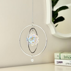 A crystal sun catcher hanging from a silver chain, featuring a faceted glass orb encased within two concentric silver rings. A small clear bead dangles from the bottom.