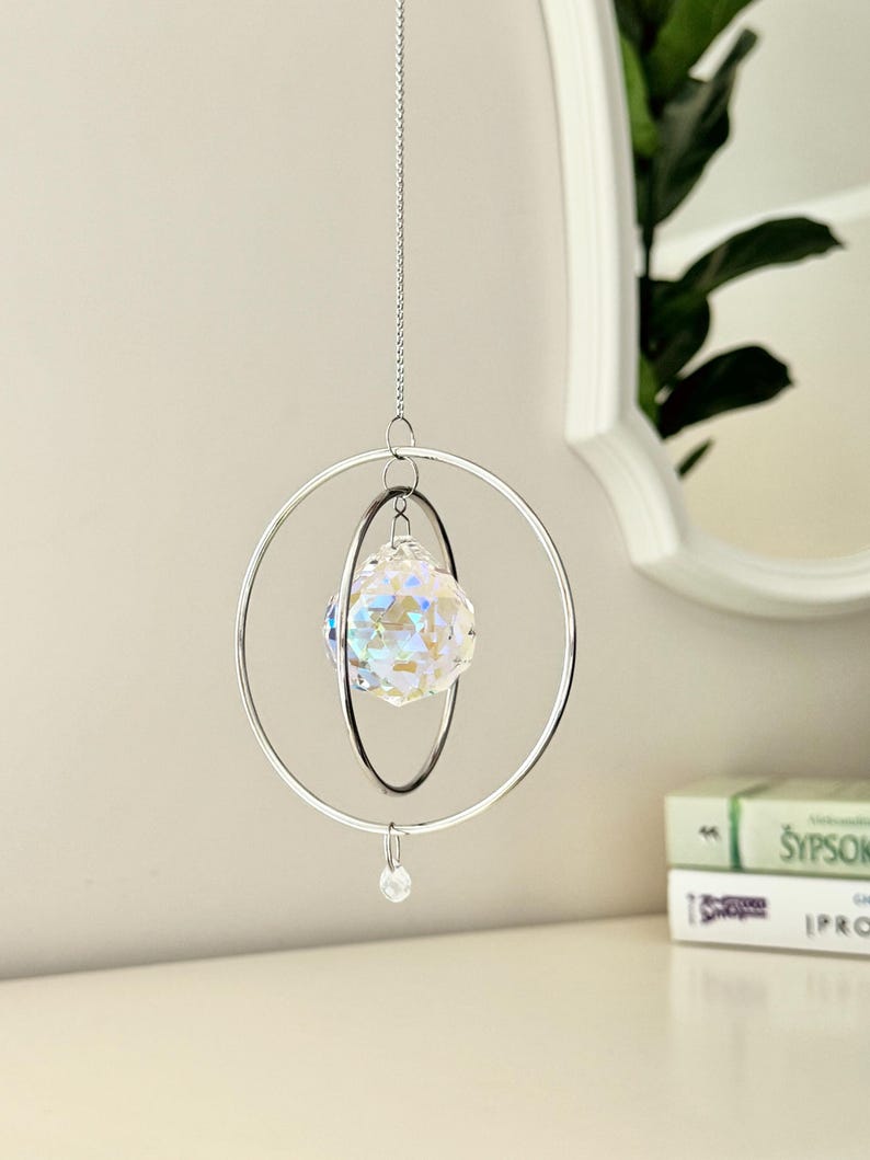 A crystal sun catcher hanging from a silver chain, featuring a faceted glass orb encased within two concentric silver rings. A small clear bead dangles from the bottom.