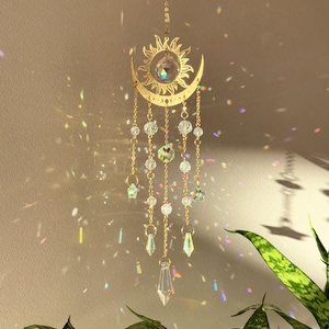 Puede incluir: Un adorno colgante dorado con forma de sol y luna creciente, con cuentas y prismas de cristal transparente. El sol tiene un gran cristal en el centro, y la luna tiene peque&ntilde;as cuentas de cristal colgando. El adorno cuelga frente a una pared.