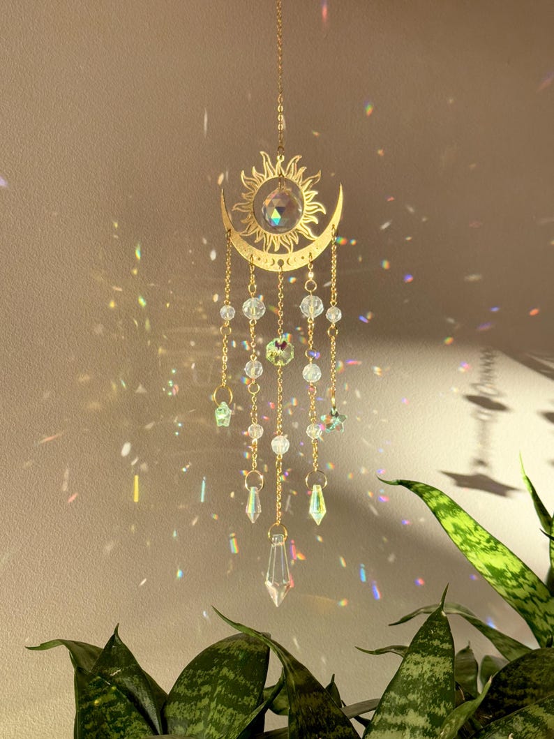 Puede incluir: Un adorno colgante dorado con forma de sol y luna creciente, con cuentas y prismas de cristal transparente. El sol tiene un gran cristal en el centro, y la luna tiene peque&ntilde;as cuentas de cristal colgando. El adorno cuelga frente a una pared.