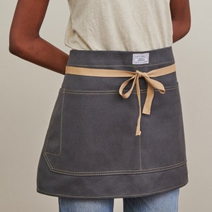 May include: A dark grey canvas apron with a tan fabric tie and a single pocket. The apron has yellow stitching around the edges and the brand name "Artifact" is printed on a white label.
