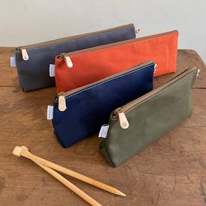 May include: Four zippered pouches made of canvas in gray, orange, navy blue, and olive green. The pouches are lined up in a row on a wooden surface. A pair of wooden knitting needles are lying on the surface.