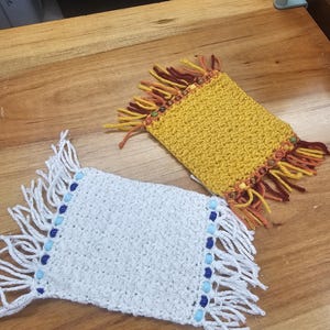 May include: Two handmade crochet coasters. One is white with blue beads and fringe, the other is yellow with brown and orange fringe. The coasters are square and appear to be made of yarn.