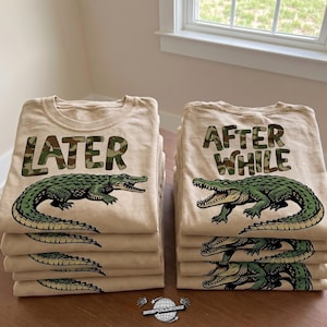 Peut inclure: Plusieurs piles de t-shirts beiges. Chaque t-shirt présente un motif d'alligator vert et un texte en camouflage. L'un dit "LATER", l'autre "AFTER WHILE". Les t-shirts sont pliés et empilés sur une surface en bois.