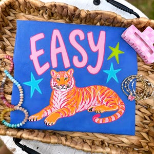 Może przedstawiać: Niebieska koszulka z napisem "EASY" w różowych literach i ilustracją tygrysa. Tygrys jest pomarańczowy z różowymi paskami. Koszulka posiada również zielone i niebieskie gwiazdki. Akcesoria obejmują bransoletki i różową spinkę do włosów.