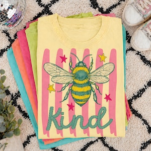 Puede incluir: Una pila de camisetas coloridas, con una camiseta amarilla en la parte superior con una gran ilustración de abeja con la palabra "Kind". La abeja es verde y amarilla, con rayas verticales rosas y acentos de estrellas. Las otras camisetas son rosas, naranjas, verdes y azules.