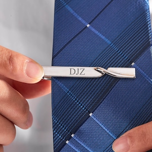 Può includere: Un fermacravatta argentato con le iniziali "DJZ" incise è agganciato a una cravatta blu a quadri. Il fermacravatta ha un design curvo e viene tenuto in posizione.