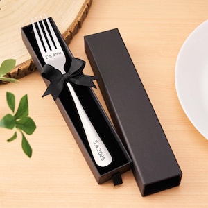 Peut inclure: Une fourchette argentée gravée avec "I'm done" et "5.4.2025" dans un coffret noir noué d'un ruban noir. La fourchette est présentée sur une surface en bois clair avec une assiette blanche en arrière-plan.