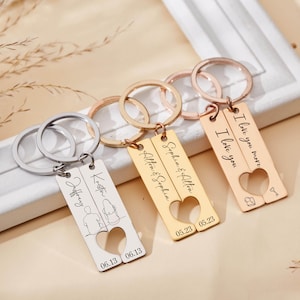 Peut inclure: Trois porte-clés personnalisés avec des découpes en forme de cœur. Un porte-clés argenté avec des noms et une date, un autre porte-clés doré avec des noms et une date, et un porte-clés or rose avec le texte "I love you more" et "I love you".