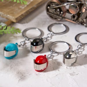 Könnte beinhalten: Schlüsselanhänger mit Miniatur-Motorradhelm-Designs in verschiedenen Farben: Türkis, Schwarz, Rot und Silber. Jeder Helm hat ein silbernes Visier und einen Schlüsselring. Einige Helme sind mit Namen graviert. Eine Motorradskulptur ist im Hintergrund.